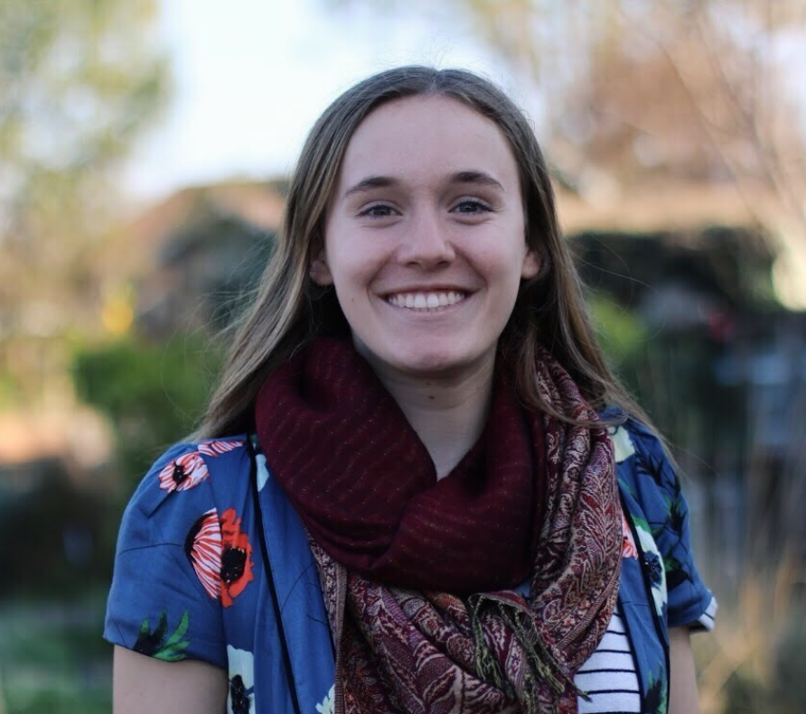 A person smiling outdoors wearing a scarf and a blue shirt.