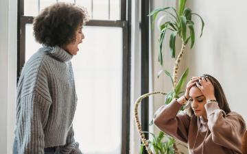 A Black woman standing at desk shouting at a seated female who appears frustrated, has hands on her head and a sad expression. Image by Liza Summer via Pexels.