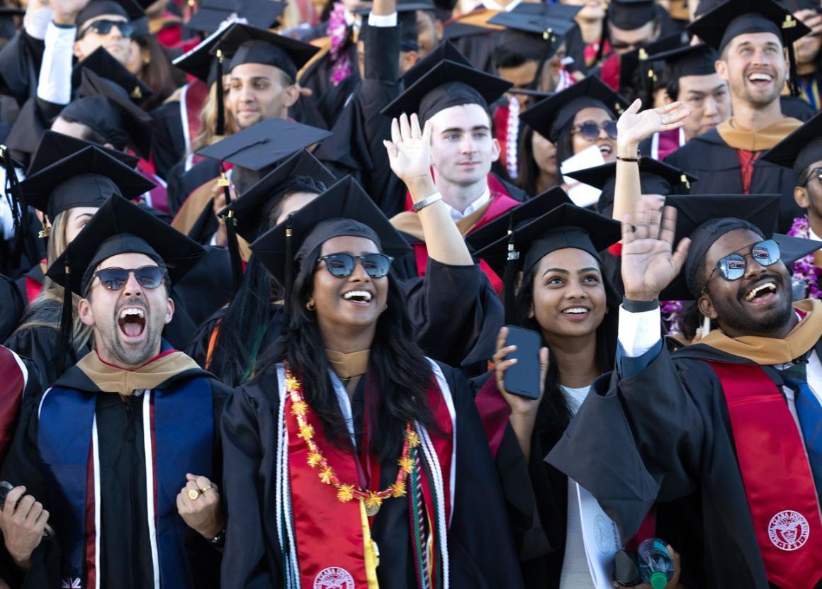 Group of graduates in caps and gowns celebrating, smiling, and waving.