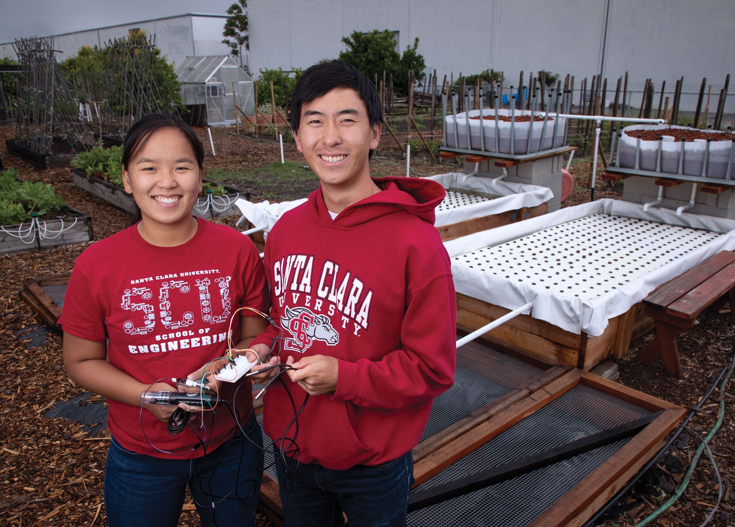 Two Santa Clara University students in red SCU apparel hold equipment in an aquaponics garden setup.