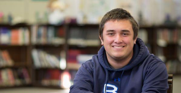 A person smiling in a library wearing a blue hoodie.