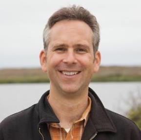 A person smiling outdoors near a body of water.