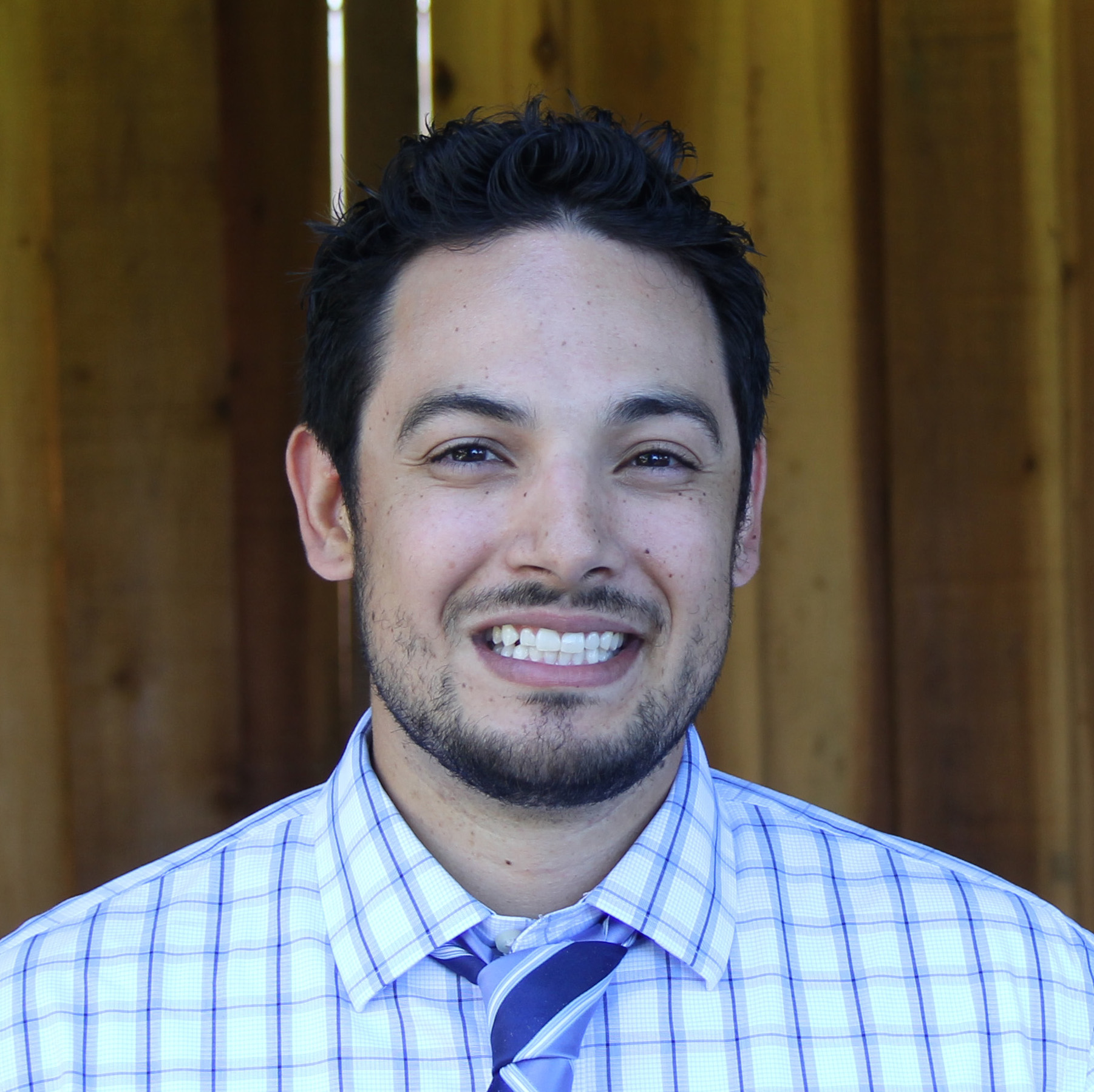 A person smiling, wearing a checkered shirt and tie.