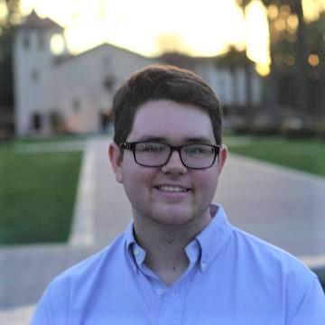 A person smiling outdoors wearing glasses and a blue shirt.