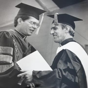 Two people wearing graduation gowns and caps, shaking hands at a commencement ceremony.