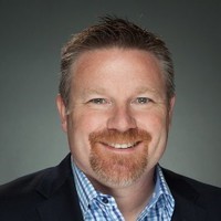 A man with a beard wearing a suit and checkered shirt, smiling.