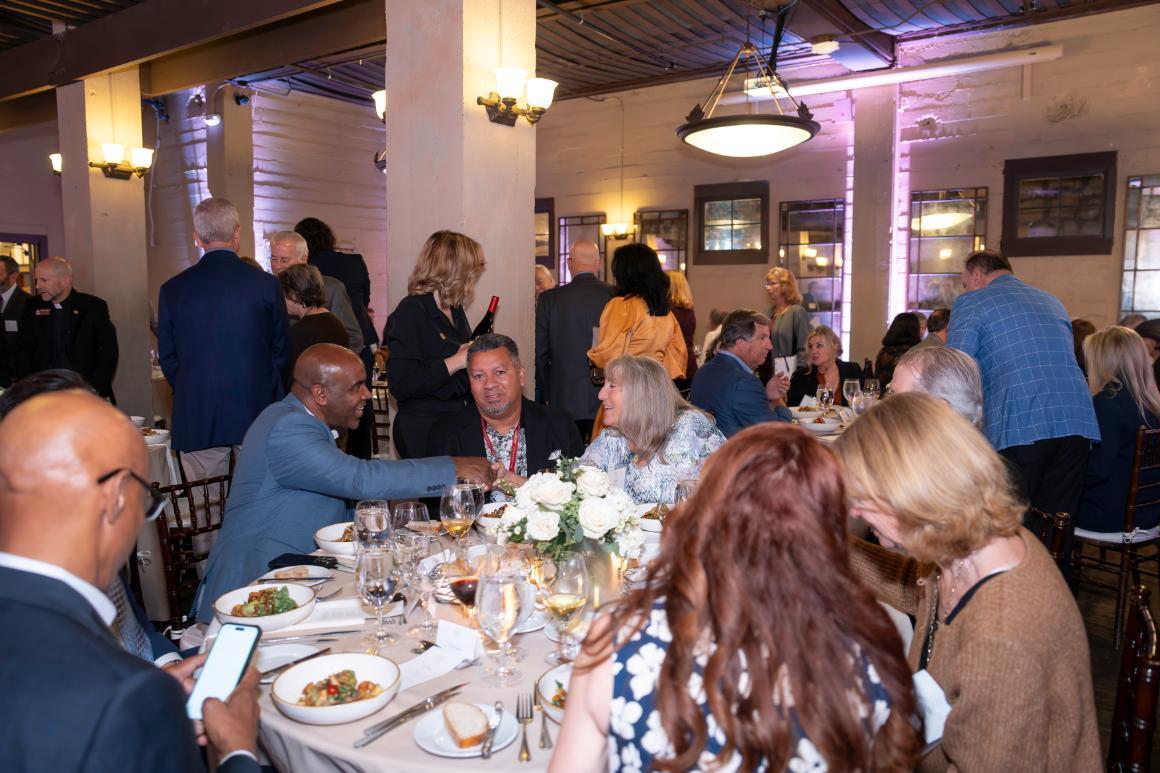 A group of diners smile and interact over a meal, while others sit at tables and mill about in the background