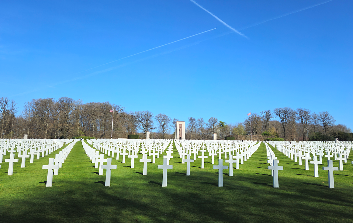 Thousands of white crosses stand on a tiny cemetery lawn with a tall stone monument in the far distance.