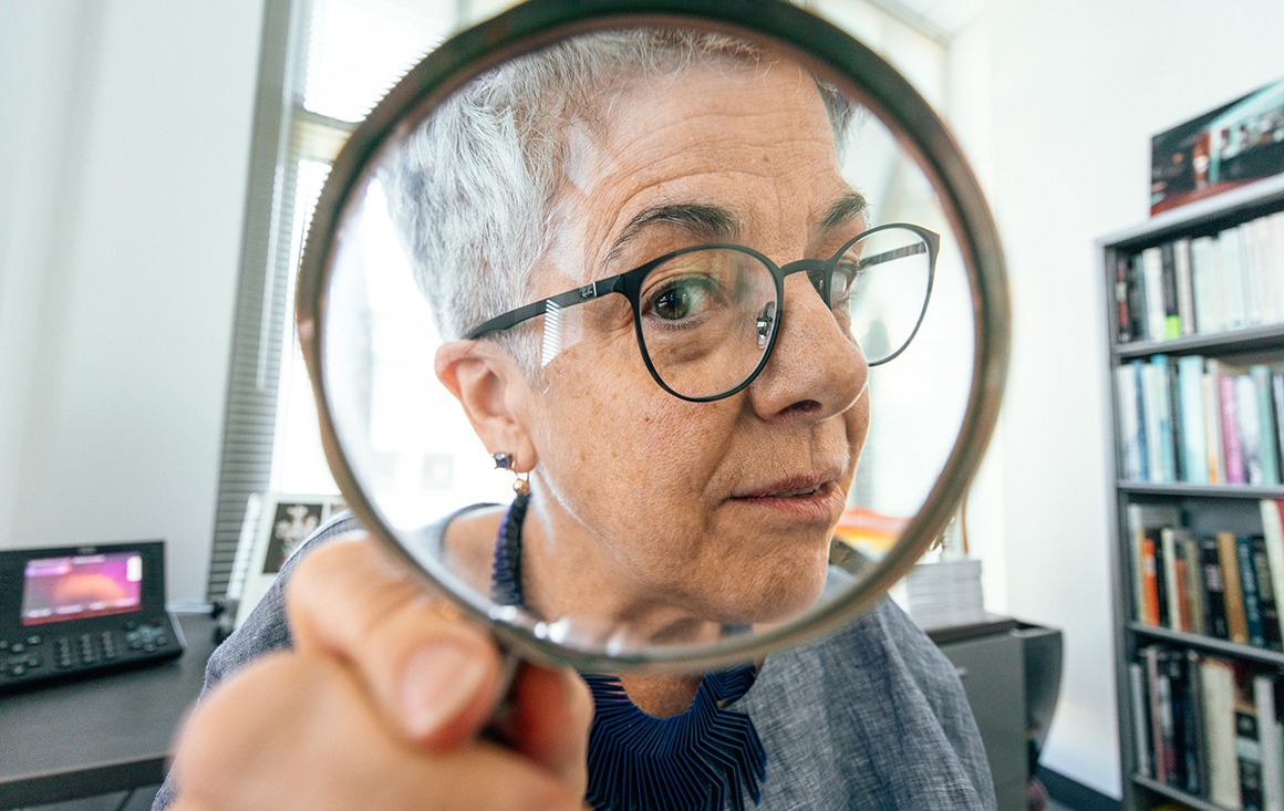 A woman with gray hair and glasses looking into the camera through a magnifying glass.