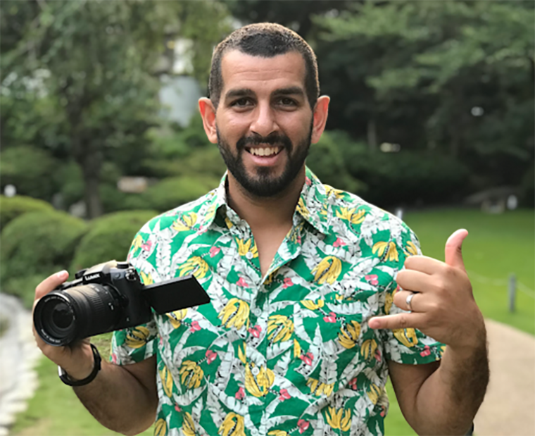 A man in a banana-patterned button up holds a camera in one hand and makes the shaka sign with the other.
