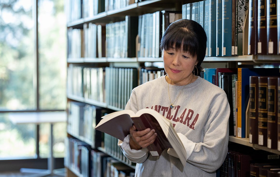 A woman stands in a library, holding a large book open to read.