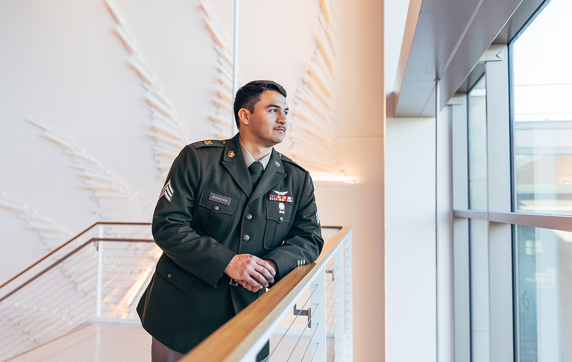 A young man in a dark green military uniform leans on a staircase handrail, looking pensively out a window.