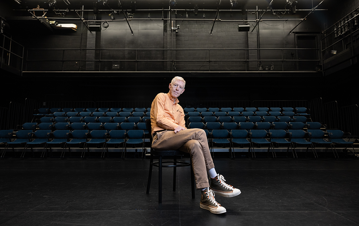 A man in an orange shirt and green sneakers sits on a chair in an empty black box theater with the seats behind him.