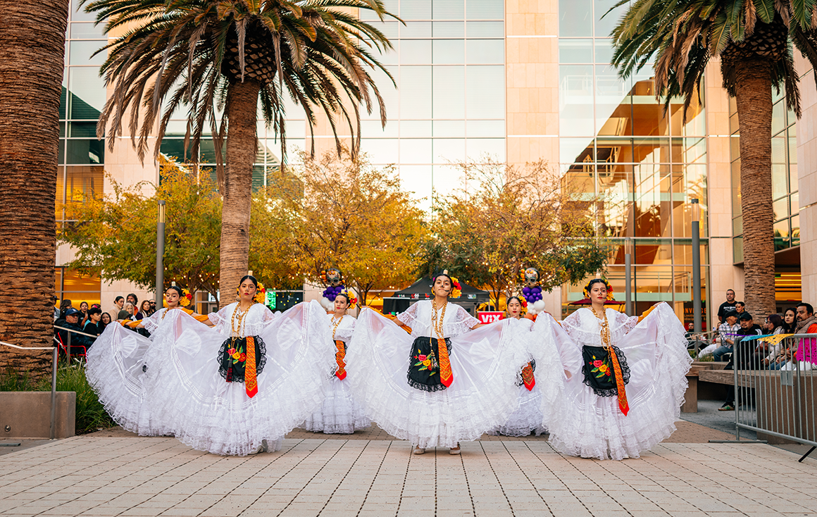 A line of women in skeleton makeup and lace white dresses with black and floral aprons stand, skirts raised in a dramatic arc, preparing to dance.