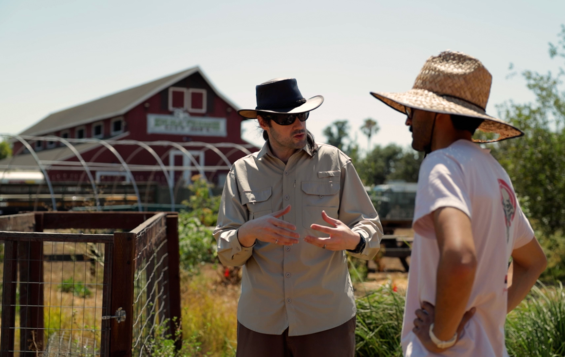 A man in a khaki shirt, leather wide-brimmed hat, and sunglasses talks emphatically with a man in a straw hat on a farm.