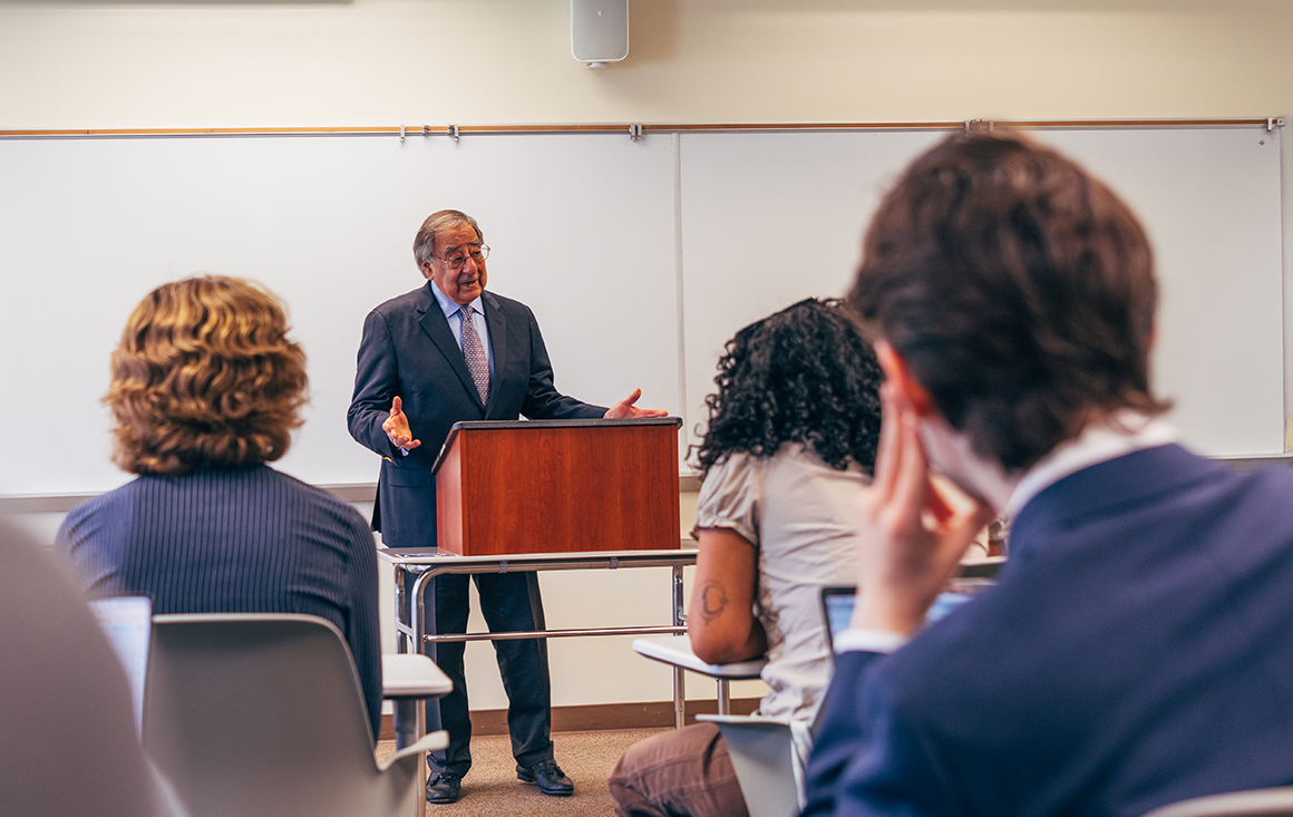 An older man lectures in front of a class of college students.
