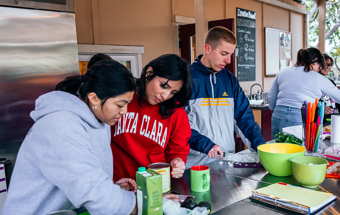 Four women and a man prepare food in an outdoor kitchen.