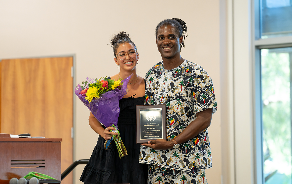 A Black student holding a bouquet of flowers poses with a Black man who is handing her an award.