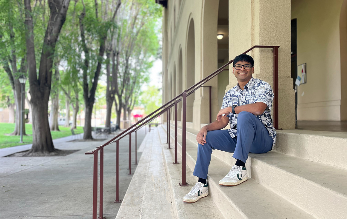 A young man in a patterned button-up and jeans sits causally on the steps to an academic building.