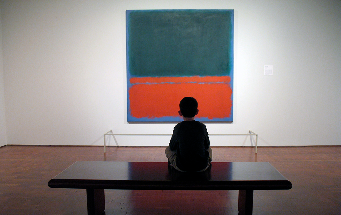 A small boy sits cross-legged on a bench in an art gallery looking at a Rothko painting consisting of a blue color block on top of orange color block.