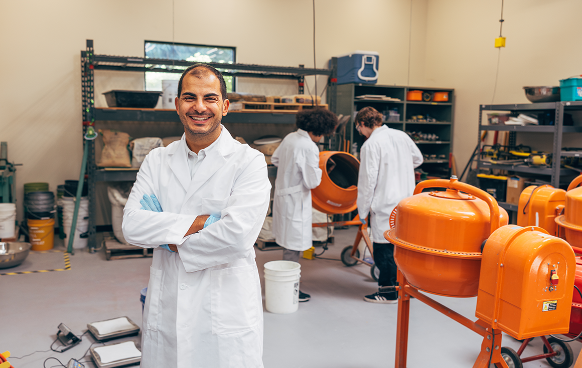 A man stands in a lab coat crossing his arms in front of two students working with a bright orange cement mixer.
