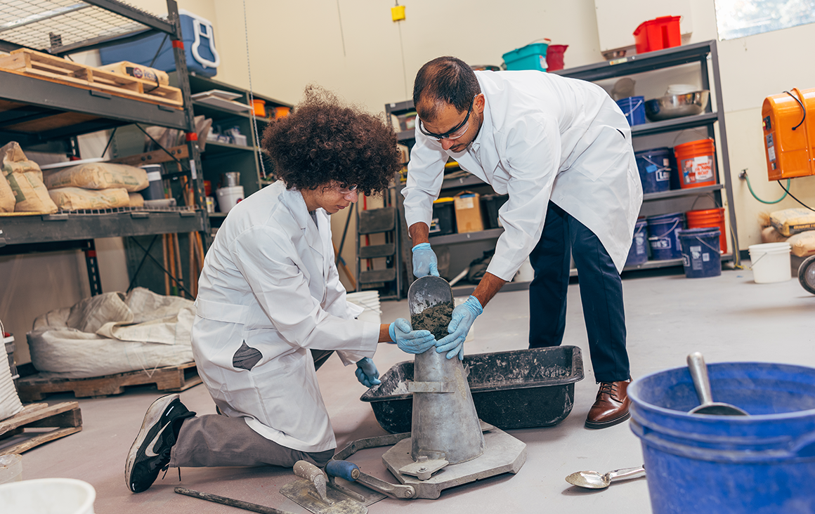 A man pours a shovel of wet cement into a metal mold being held by a student. Both are wearing lab coats in an industrial space.