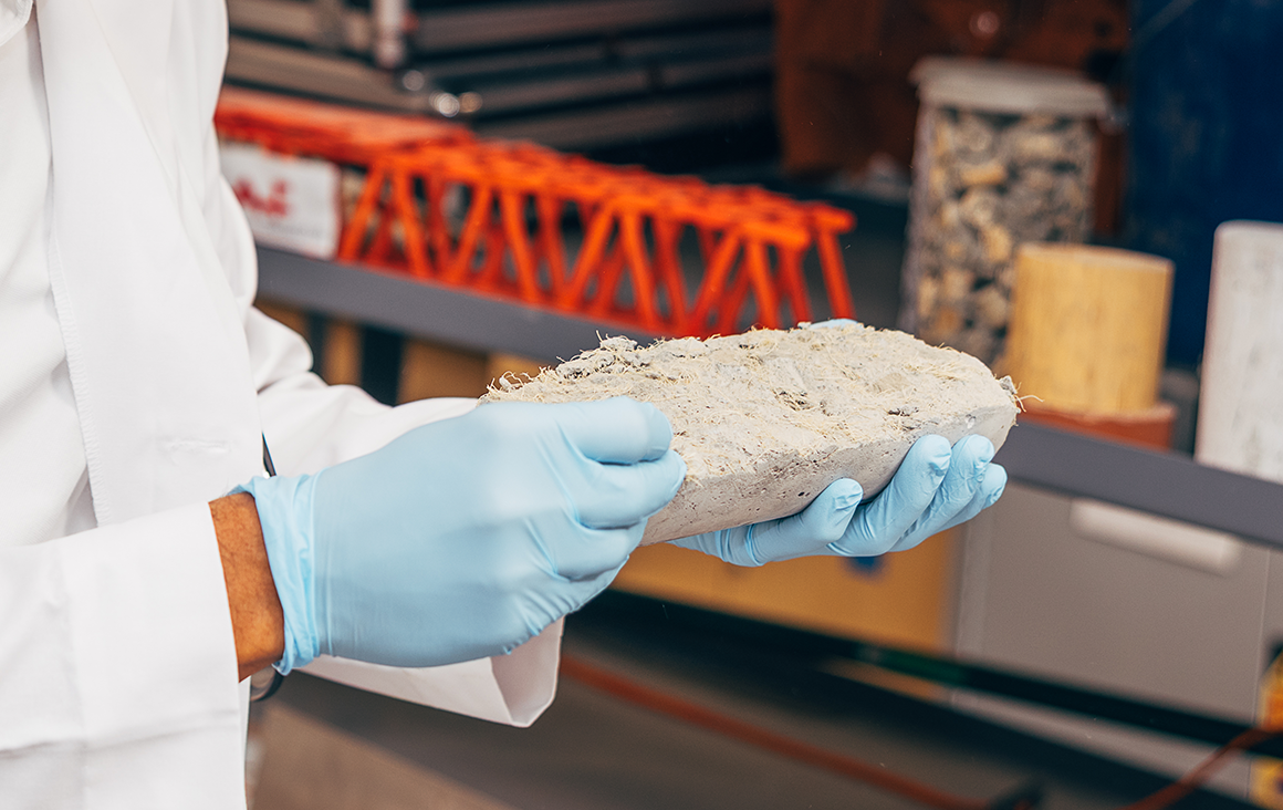 A man wearing blue nitrile gloves holds a cylinder of cement, split in half vertically, revealing plant fibers inside.