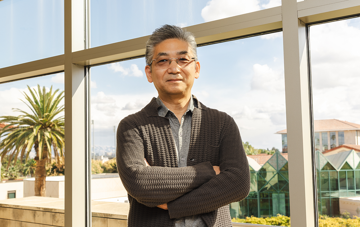 A Korean man in tinted glasses and a brown cardigan stands, arms crossed, in front of a window, palm tree and SCU campus behind him.