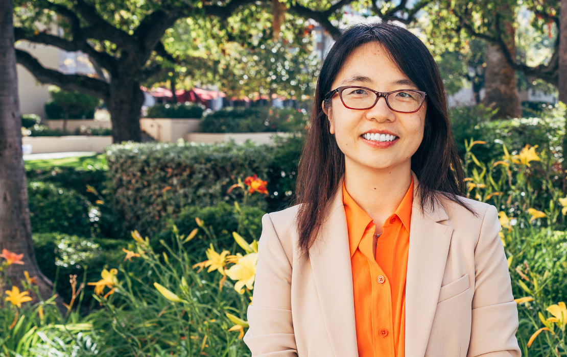 a woman in a jacket and blouse is standing in front of flowers and trees