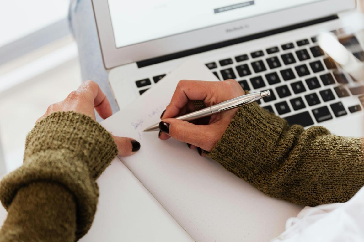 Hands holding a pen, writing on notebook next to an open laptop