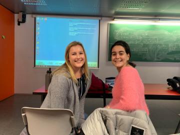 Two women smiling, seated in a classroom with a projector screen and whiteboard behind them.