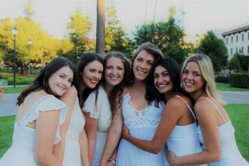 Group of six women smiling and hugging outdoors.