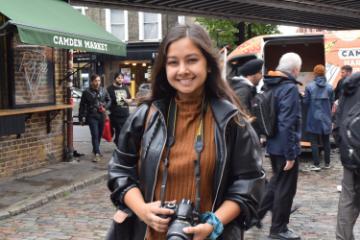 A woman in a market, smiling, wearing a brown dress and black jacket.