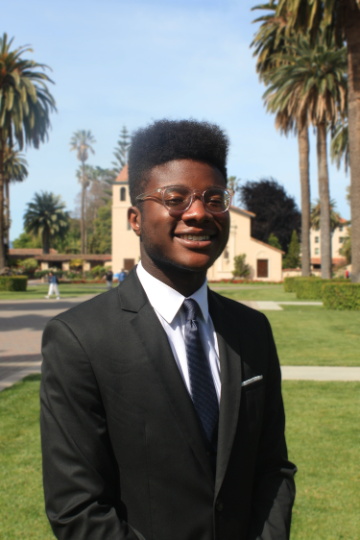 A person in a suit standing outdoors with palm trees in the background.