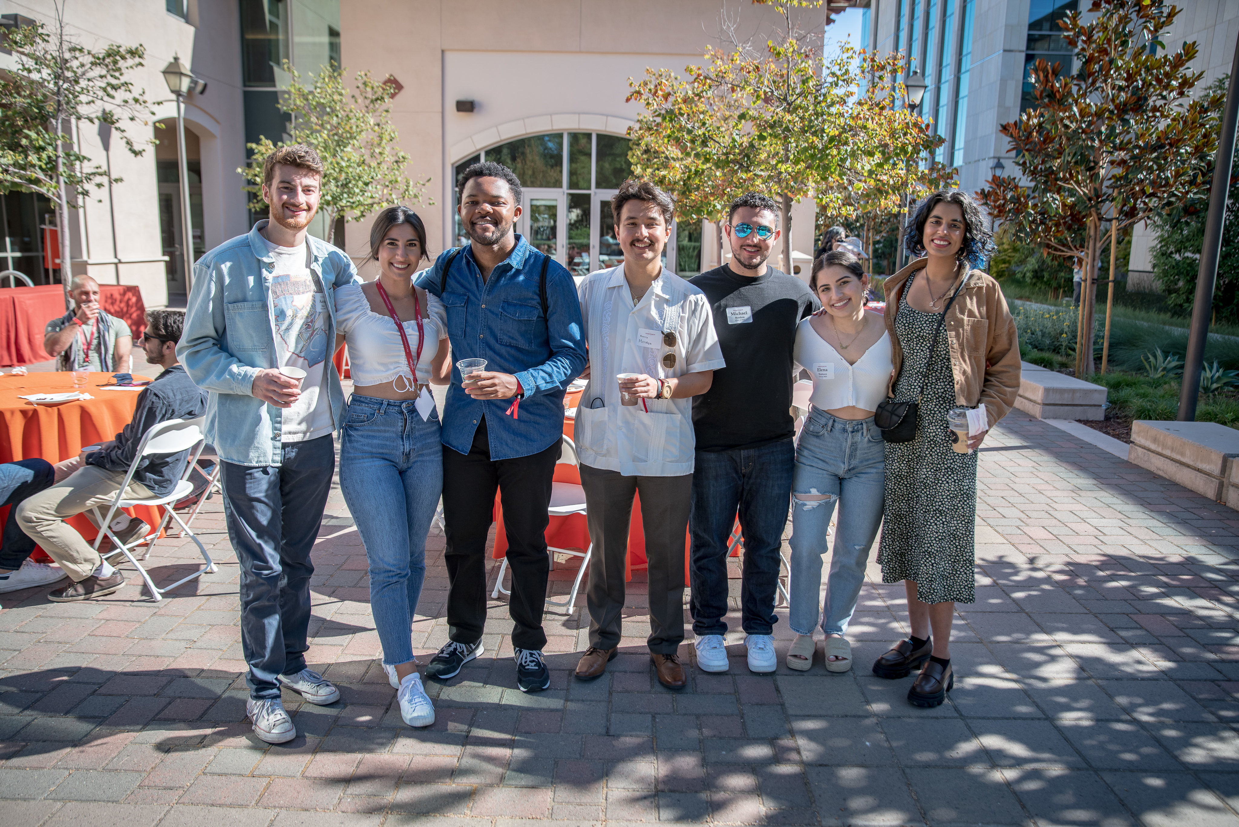 Seven alumni stand together with their arms around each other, smiling at the camera