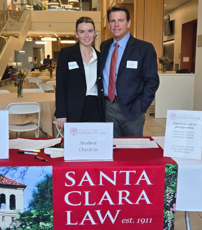 A man and a young woman in business wear standing behind an event check-in table with a banner reading 