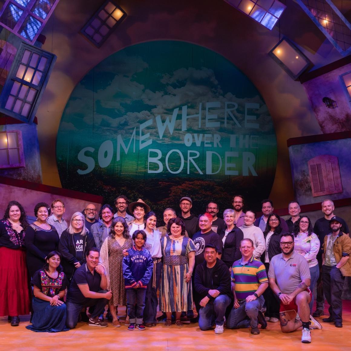 A group of people, some in costume, stand on a theater stage in front of a screen that reads Somewhere Over the Border in a whimsical font.