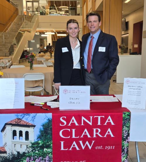 A man and a young woman in business wear standing behind an event check-in table with a banner reading
