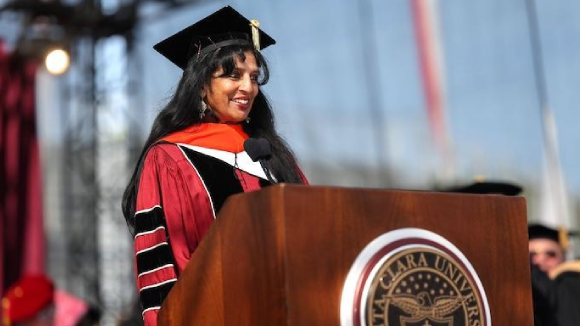 A woman in commencement regalia speaks from a podium bearing the Santa Clara University seal.