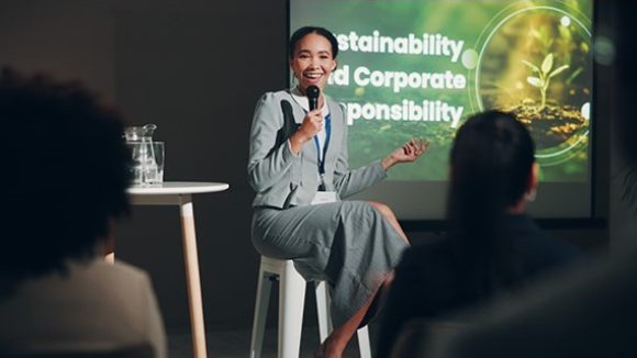 A smiling young woman seated in front of a large display screen. The text on the screen is cut off slightly by her head and torso but reads