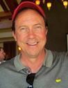 A man smiling, wearing a name tag and sunglasses hanging from his shirt.