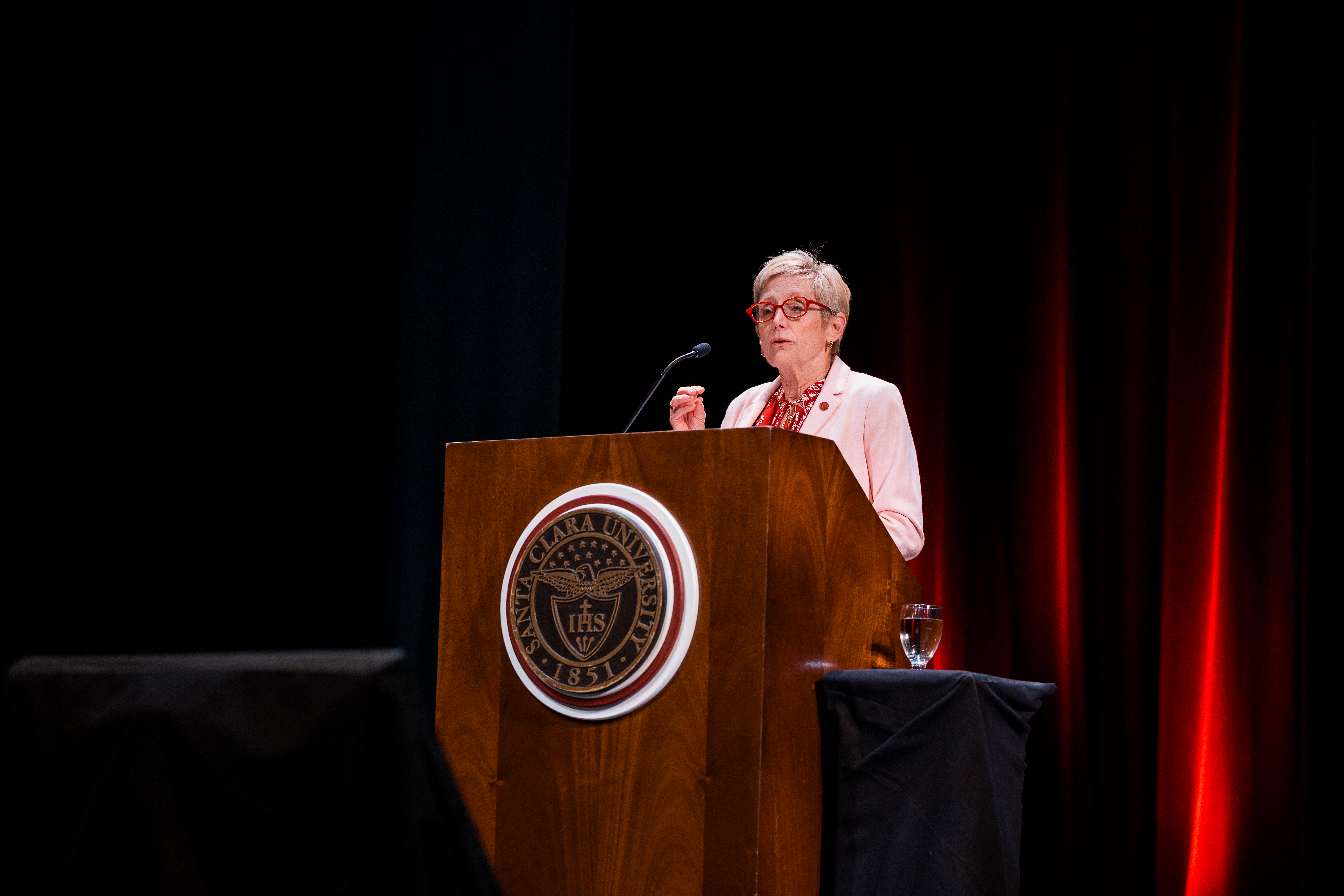 Santa Clara University President Julie Sullivan speaks at a podium on stage, wearing a light pink blazer and red glasses.