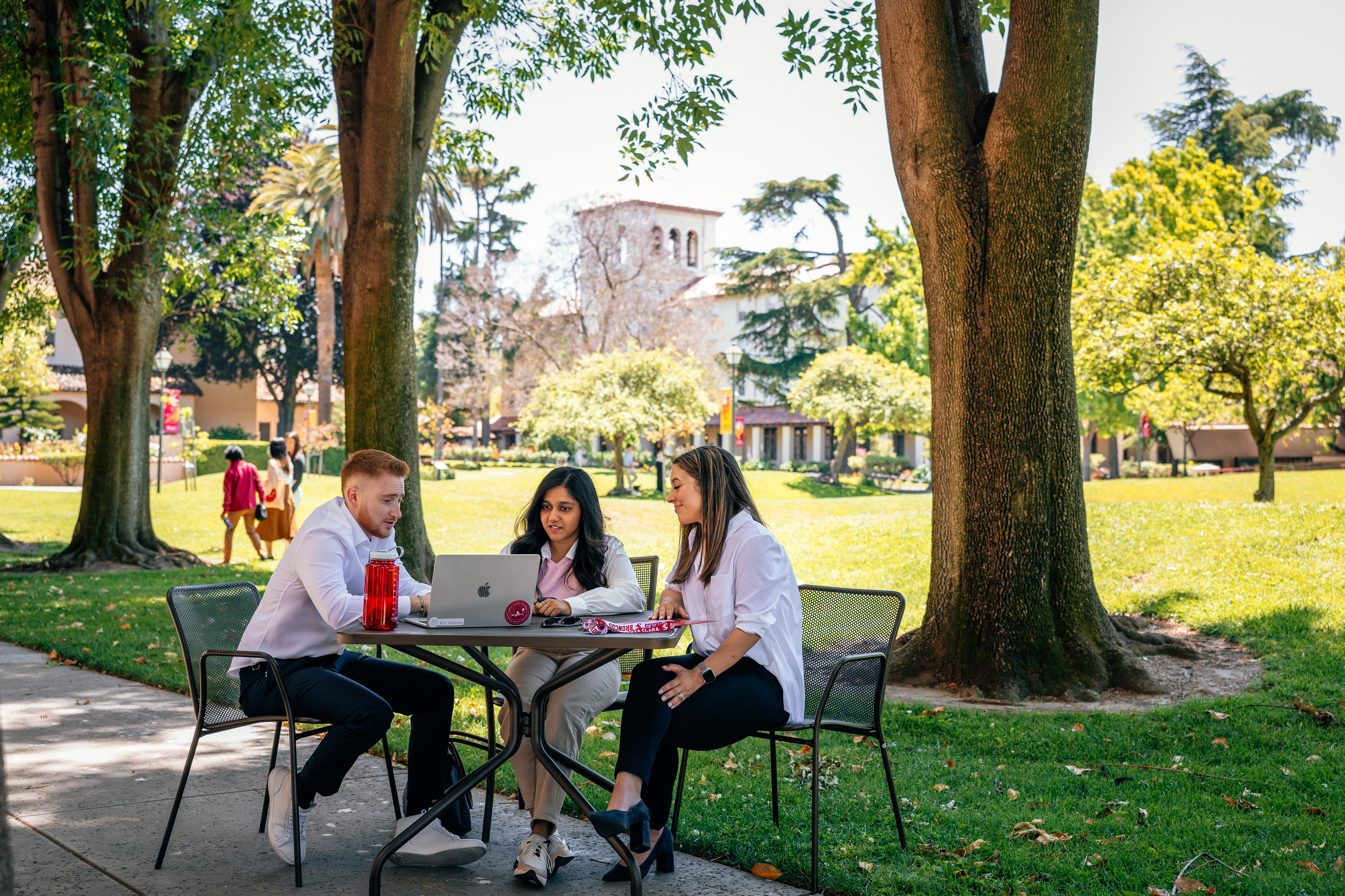 Three people sitting at a table with laptops outside 