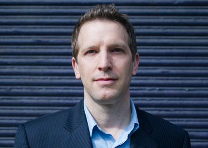 Alt text: Close-up of a man in a suit jacket against a dark striped background.