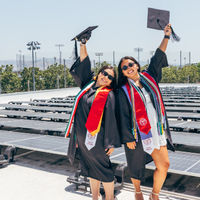 Photo of two scu seniors in graduation gowns posing in from the Bird Solar panels atop AEC holding their caps up