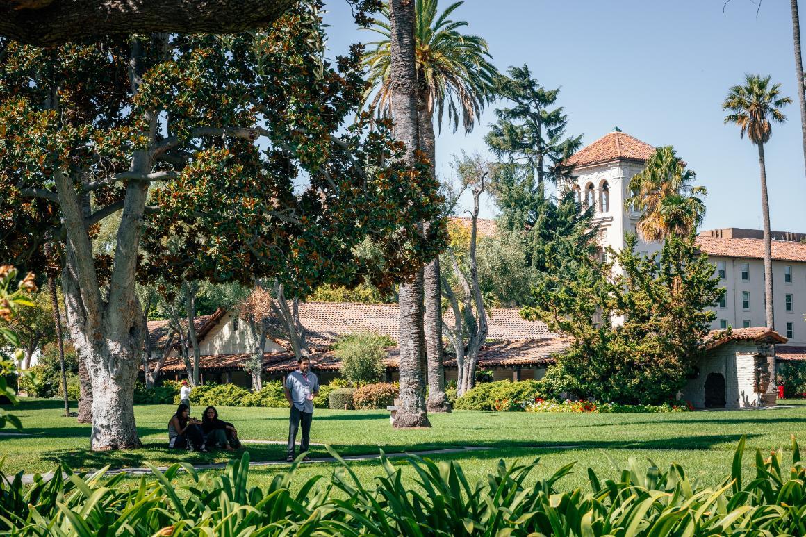 People sitting and standing under a tree on the lawn near the Adobe Lodge