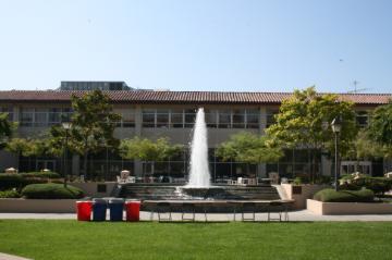 The image shows a large building with a central fountain and green lawn surrounded by trees.