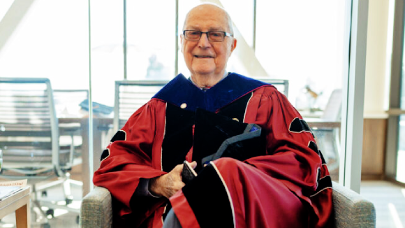 An older man in glasses sitting down, wearing full commencement regalia