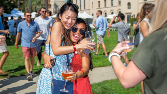 Two women smiling and laughing with wine glasses in hand while another woman takes a picture of them.