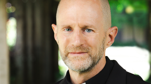 A White man in a black shirt with a white clerical collar